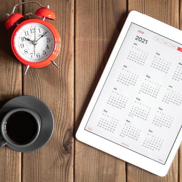 tablet with calendar for 2021, a cup of coffee and a red alarm clock on a wooden background flat lay